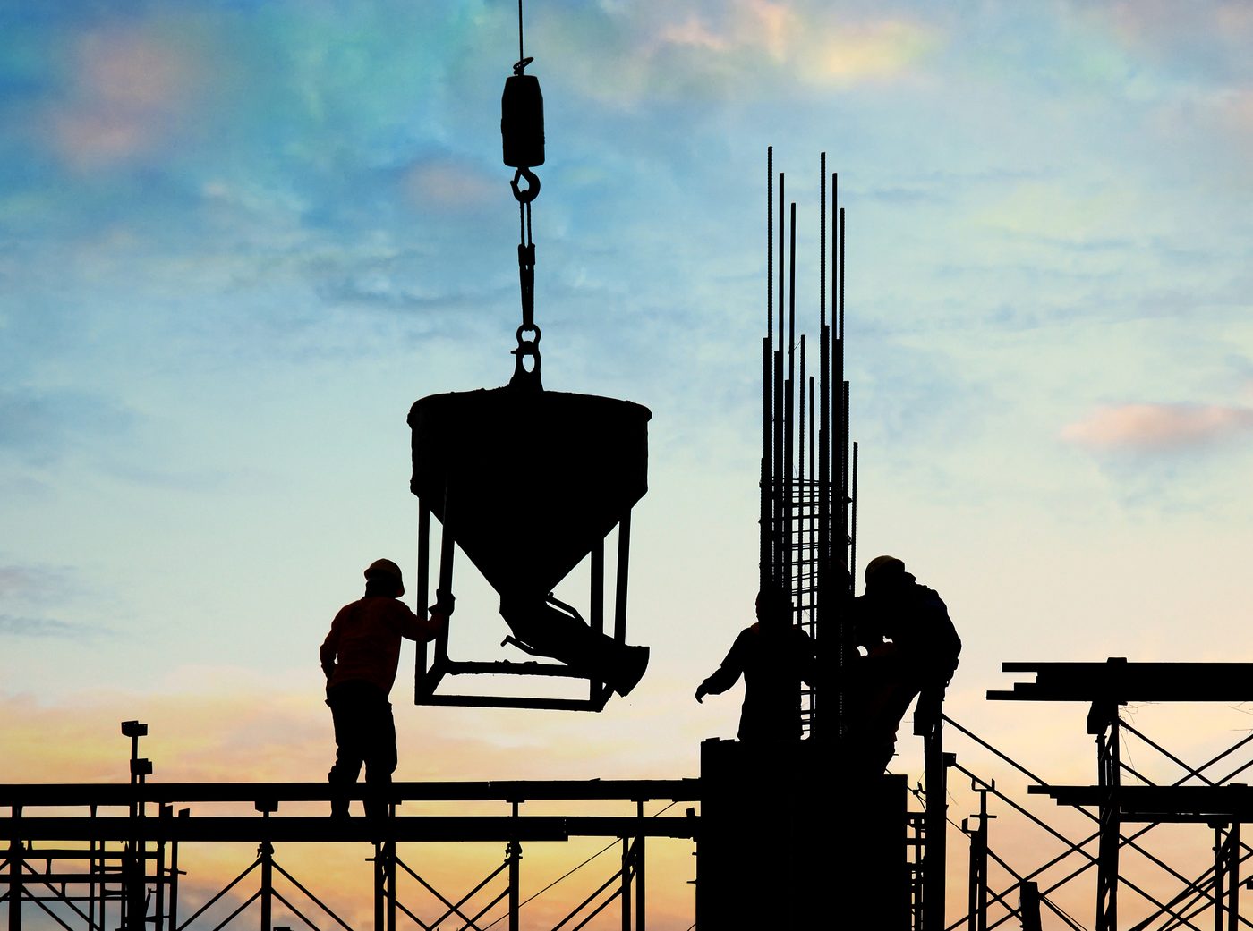 Silueta de los maestros levantando con la grúa una cubeta de concreto, sobre la estructura de la obra, al atardecer