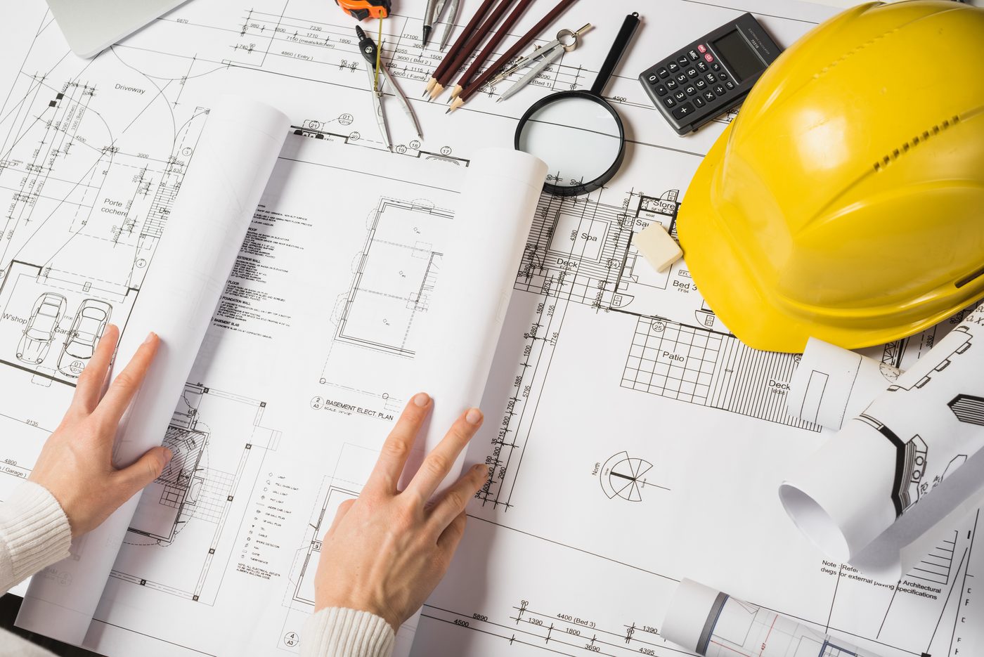 Manos revisando los planos de una vivienda, junto al casco de obra, el compás y la calculadora